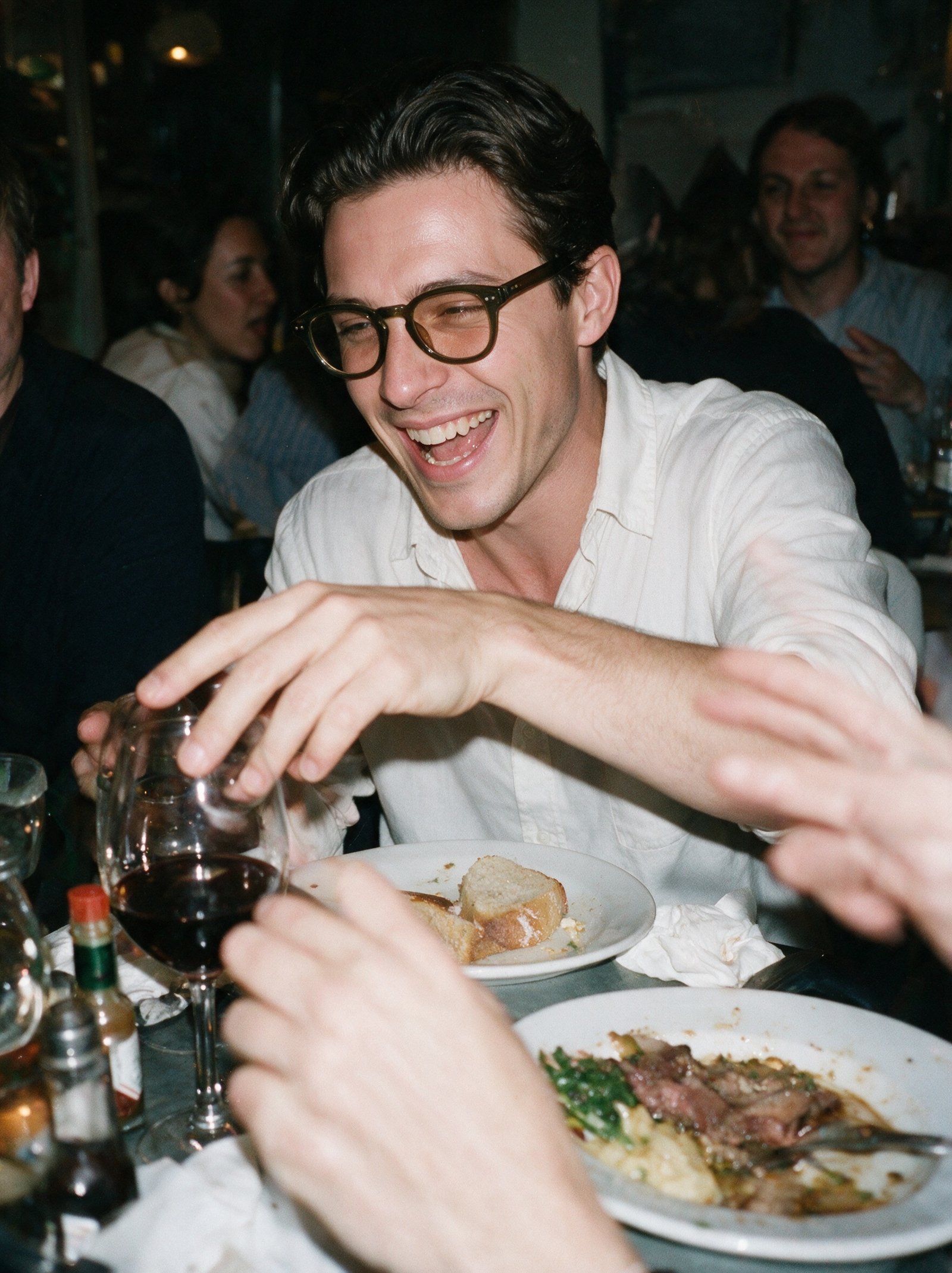 Man laughing at a crowded restaurant table, wearing the olive Bohemian frame